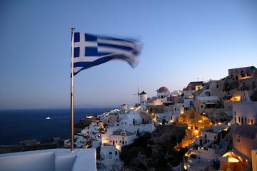 A Greek flag waving in the foreground overlooking the white-washed, cliffside buildings of a coastal village at dusk, with the sea and a distant ship in the background.
