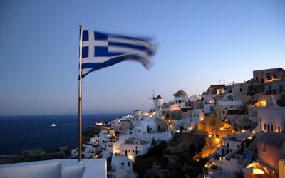 A Greek flag waving in the foreground overlooking the white-washed, cliffside buildings of a coastal village at dusk, with the sea and a distant ship in the background.