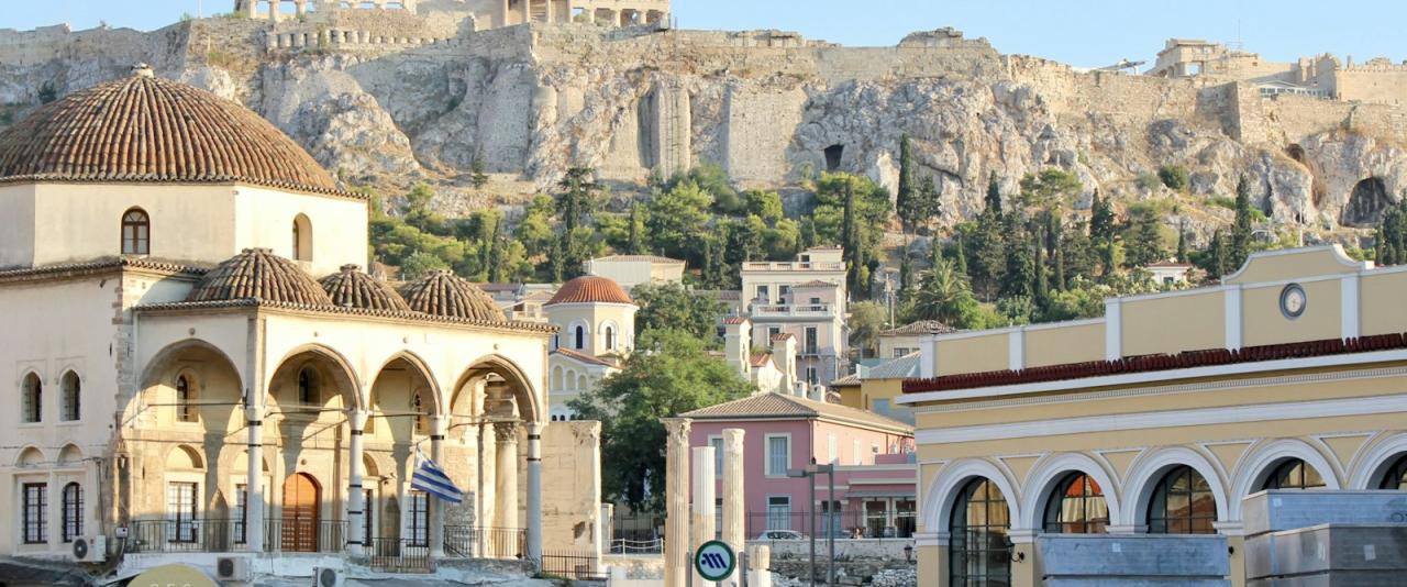 A busy street scene in Athens featuring the Byzantine church of the Metamorphosis Sotira in the foreground with the Acropolis and Parthenon visible on the hill in the background.