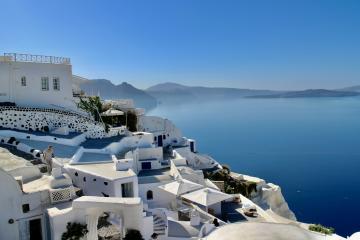 A view of white-washed cubic buildings and staircases cascading down a cliffside overlooking the deep blue Aegean Sea with distant volcanic islands in the background.