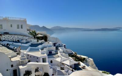 A view of white-washed cubic buildings and staircases cascading down a cliffside overlooking the deep blue Aegean Sea with distant volcanic islands in the background.