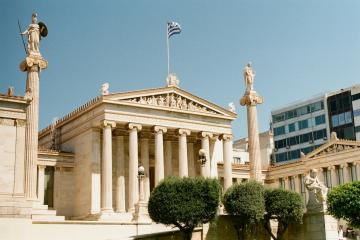 The image shows the facade of the Academy of Athens, a neoclassical building featuring a portico with columns, statues on tall pillars, and a Greek flag flying above.