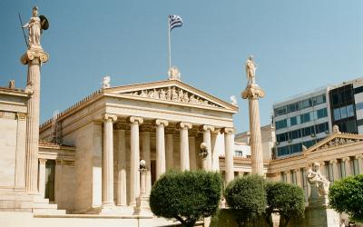 The image shows the facade of the Academy of Athens, a neoclassical building featuring a portico with columns, statues on tall pillars, and a Greek flag flying above.