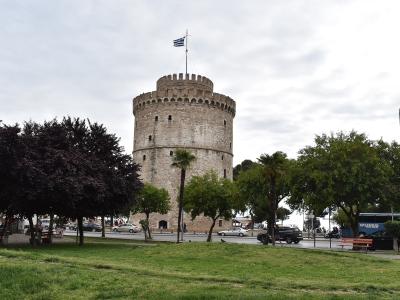 A large, cylindrical stone tower with crenellations and a Greek flag flying from the top, situated next to a road with cars and trees in a park-like setting.