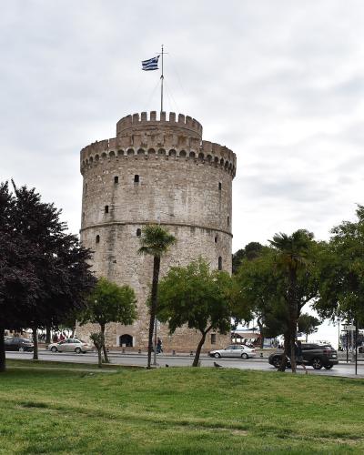 A large, cylindrical stone tower with crenellations and a Greek flag flying from the top, situated next to a road with cars and trees in a park-like setting.