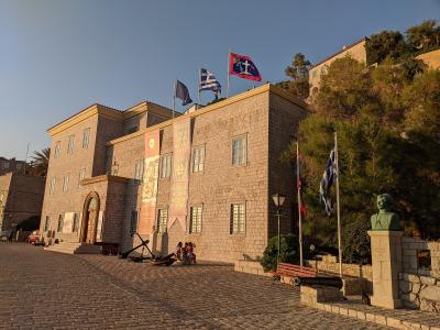 A large stone building with flags on the roof and banners hanging from the facade, situated on a cobblestone street with a bust statue nearby.