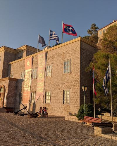 A large stone building with flags on the roof and banners hanging from the facade, situated on a cobblestone street with a bust statue nearby.