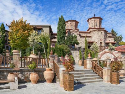 A stone-paved courtyard with large terracotta pots, wooden railings, and a stone church building with two domes in the background under a blue sky.