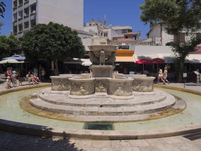 A large, ornate stone fountain featuring a central basin supported by winged lions, situated in a paved public square with surrounding cafes and buildings.