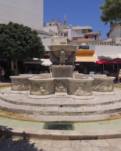 A large, ornate stone fountain featuring a central basin supported by winged lions, situated in a paved public square with surrounding cafes and buildings.