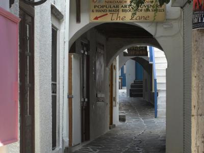 A narrow, stone-paved alleyway in a Greek town featuring whitewashed walls, an archway, and shop signs including one for "Glamour Handmade Jewellery" and another for "The Lane" souvenir shop with Greek text.