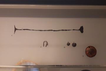 Museum display case containing ancient metal artifacts, including a long bar, a horseshoe-shaped object, small ingots, and a round coin, labeled with numbers 7, 9, 10, and 11.