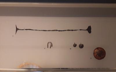 Museum display case containing ancient metal artifacts, including a long bar, a horseshoe-shaped object, small ingots, and a round coin, labeled with numbers 7, 9, 10, and 11.