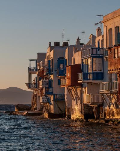 A row of traditional wooden houses built on stilts over the water at sunset, with a distant island silhouette in the background.