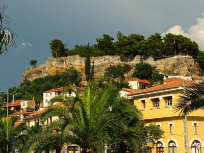 A stone fortress wall atop a hill overlooking a town with yellow buildings and palm trees, with a Greek flag flying on the right side of the hilltop.