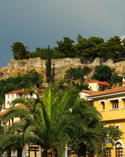 A stone fortress wall atop a hill overlooking a town with yellow buildings and palm trees, with a Greek flag flying on the right side of the hilltop.
