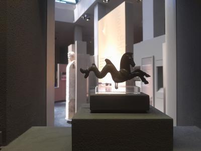 The image shows a bronze figurine of a horse with a serpent-like tail on a pedestal inside a museum, with a marble statue visible in the background.
