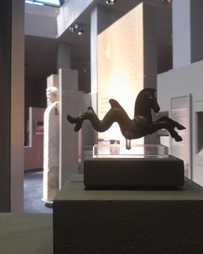 The image shows a bronze figurine of a horse with a serpent-like tail on a pedestal inside a museum, with a marble statue visible in the background.