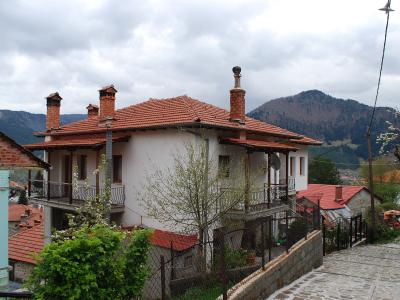 A traditional stone and stucco house with a terracotta tiled roof, wooden balcony, and multiple chimneys, situated on a sloping street with other similar buildings and mountains in the background.