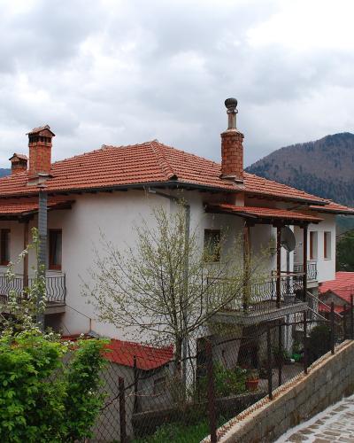 A traditional stone and stucco house with a terracotta tiled roof, wooden balcony, and multiple chimneys, situated on a sloping street with other similar buildings and mountains in the background.