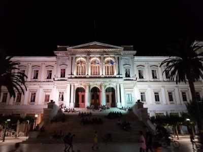 A large neoclassical building illuminated at night, featuring a central portico with columns, three arched doorways, and a wide staircase leading up to the entrance where many people are gathered.