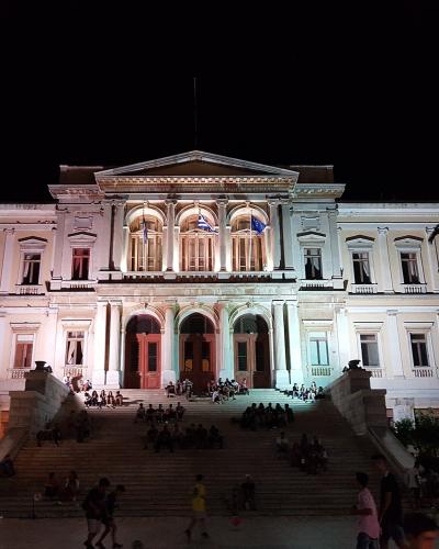 A large neoclassical building illuminated at night, featuring a central portico with columns, three arched doorways, and a wide staircase leading up to the entrance where many people are gathered.