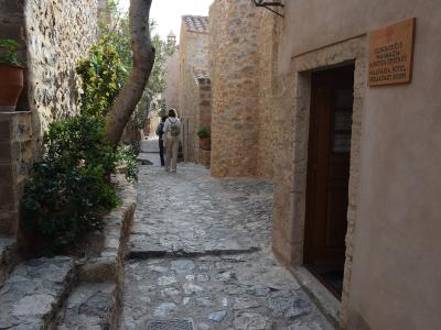 A narrow, cobblestone alleyway in a historic stone village with two people walking away, featuring a wooden sign on the right wall.