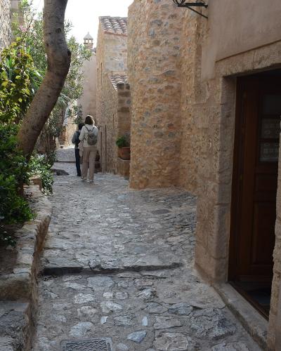 A narrow, cobblestone alleyway in a historic stone village with two people walking away, featuring a wooden sign on the right wall.