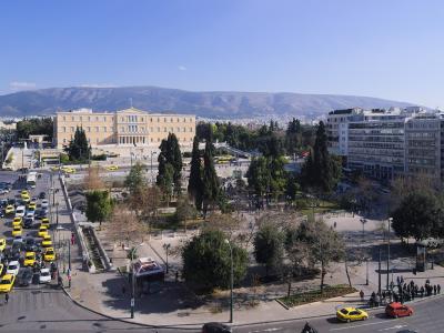 A high-angle view of a large urban square featuring a prominent neoclassical government building (the Hellenic Parliament) on the left, a central park area with trees, and busy streets with yellow taxis in the foreground.