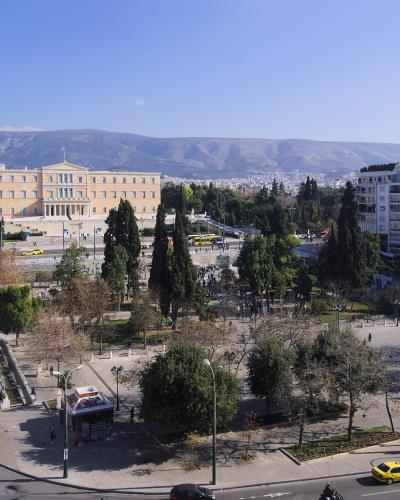 A high-angle view of a large urban square featuring a prominent neoclassical government building (the Hellenic Parliament) on the left, a central park area with trees, and busy streets with yellow taxis in the foreground.