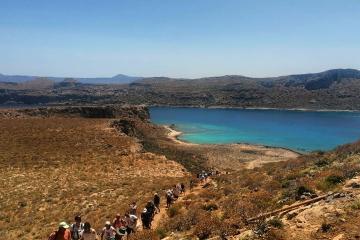 A group of hikers walking along a dirt trail on a dry, arid hillside overlooking a turquoise coastal inlet with a small sandy beach and distant hills.