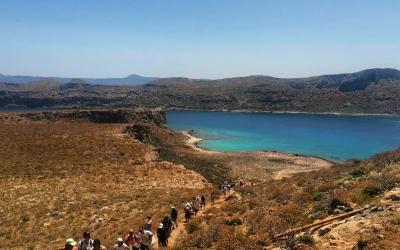 A group of hikers walking along a dirt trail on a dry, arid hillside overlooking a turquoise coastal inlet with a small sandy beach and distant hills.