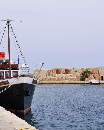 A black and white boat named "RINI" is moored at a stone pier in a harbor, with historic stone fortifications and buildings visible in the background.
