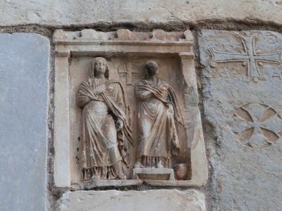 A close-up photograph of a stone relief carving on an exterior wall, depicting two standing figures within a niche, flanked by carved crosses and a frieze with birds below.