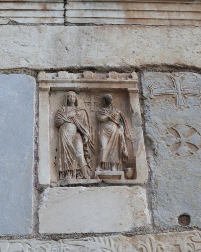 A close-up photograph of a stone relief carving on an exterior wall, depicting two standing figures within a niche, flanked by carved crosses and a frieze with birds below.