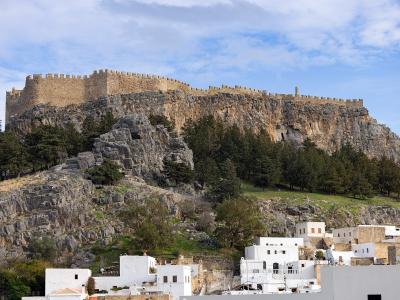 A large medieval fortress with crenellated stone walls sits atop a steep, rocky cliff, overlooking a cluster of white buildings and trees below under a blue sky.