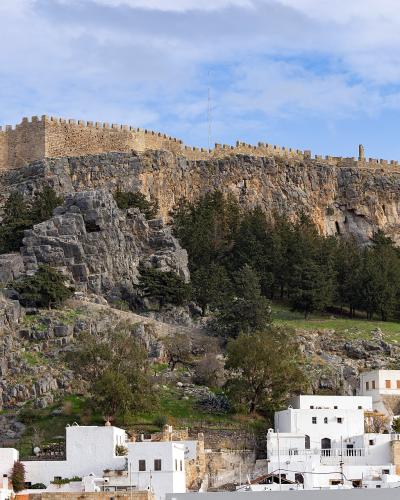 A large medieval fortress with crenellated stone walls sits atop a steep, rocky cliff, overlooking a cluster of white buildings and trees below under a blue sky.
