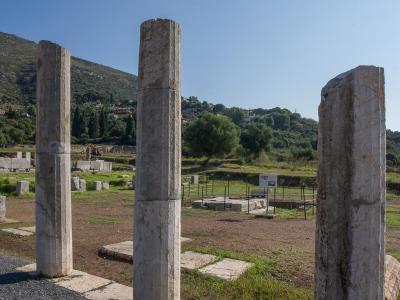 Three standing ancient stone columns in the foreground of an archaeological excavation site with ruins and a hillside village in the background.