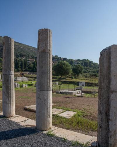 Three standing ancient stone columns in the foreground of an archaeological excavation site with ruins and a hillside village in the background.