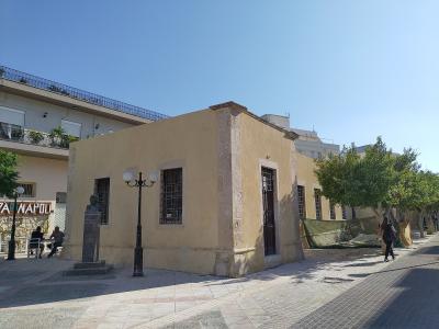 A single-story, weathered yellow building with barred windows and a bust on a pedestal in front, situated on a paved pedestrian street.
