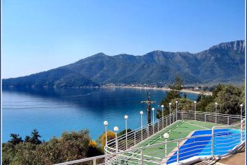 A view from an elevated terrace with white railings and blue-and-green painted steps overlooking a bay with a sandy beach and forested mountains in the background.