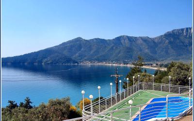 A view from an elevated terrace with white railings and blue-and-green painted steps overlooking a bay with a sandy beach and forested mountains in the background.