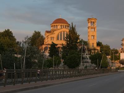 A large Byzantine-style church with a central dome and a separate bell tower flying a Greek flag, situated on a hill overlooking a road and bridge.