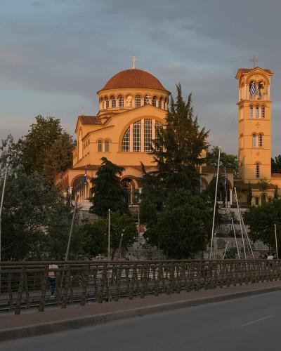A large Byzantine-style church with a central dome and a separate bell tower flying a Greek flag, situated on a hill overlooking a road and bridge.