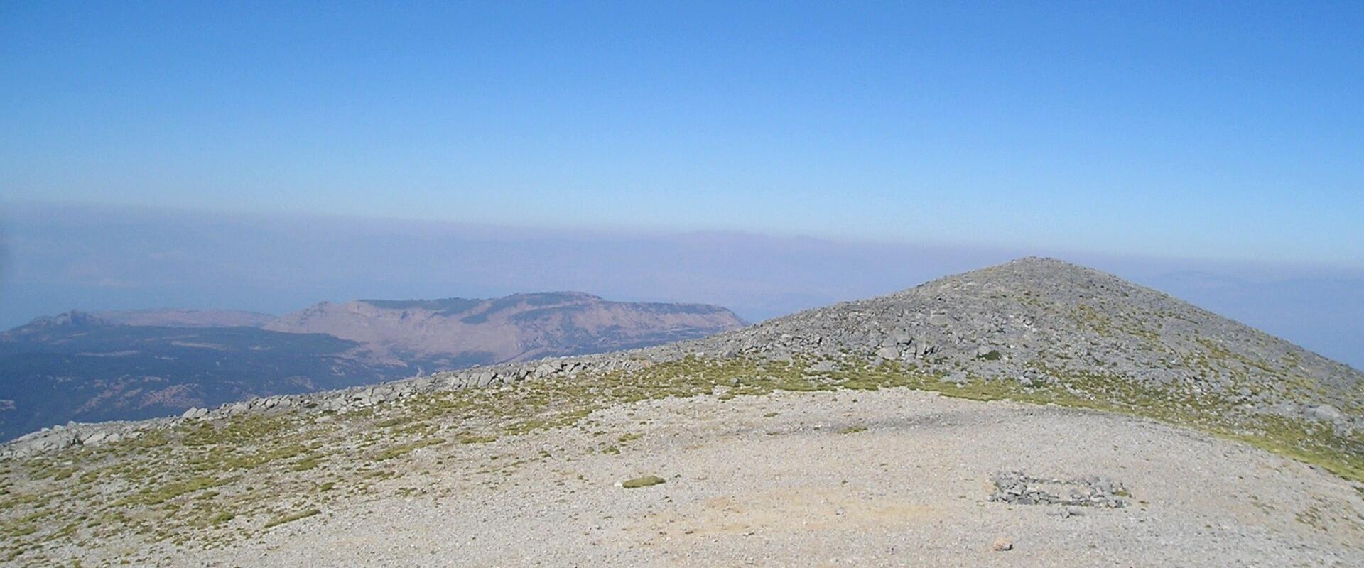 A rocky, gravelly mountain summit with sparse vegetation under a clear blue sky, featuring a low stone cairn or marker in the foreground and distant mountain ridges on the horizon.