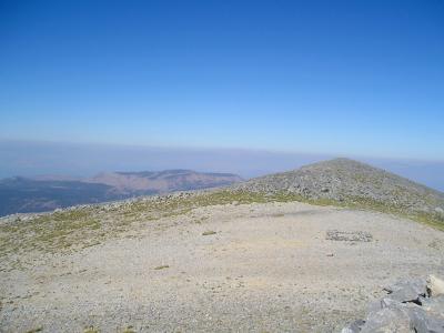 A rocky, gravelly mountain summit with sparse vegetation under a clear blue sky, featuring a low stone cairn or marker in the foreground and distant mountain ridges on the horizon.