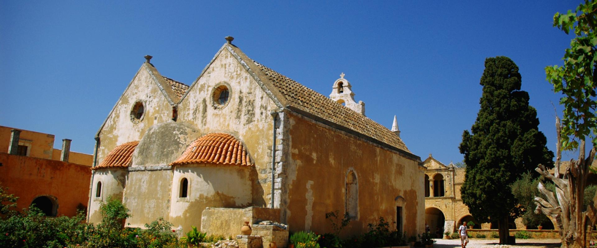 A weathered, ochre-colored stone monastery complex with a terracotta-tiled roof, a small bell tower, and arched windows, set against a clear blue sky with a large cypress tree to the right.