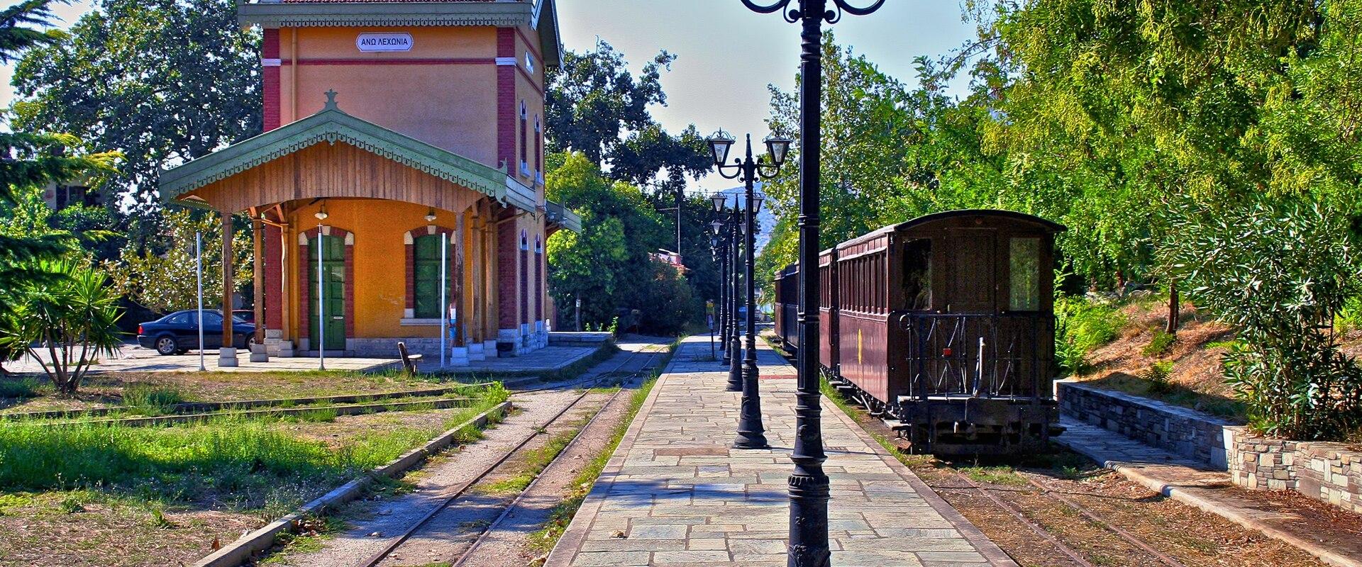 A narrow-gauge railway station building with a yellow facade and red roof, a vintage wooden train car on the tracks, and a stone platform lined with black lampposts.