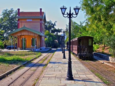 A narrow-gauge railway station building with a yellow facade and red roof, a vintage wooden train car on the tracks, and a stone platform lined with black lampposts.