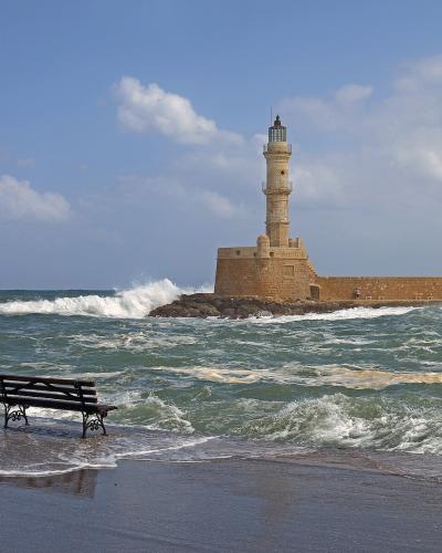 A stone lighthouse situated at the end of a long harbor breakwater, viewed from a wet promenade with a bench and lamp post in the foreground during rough seas.